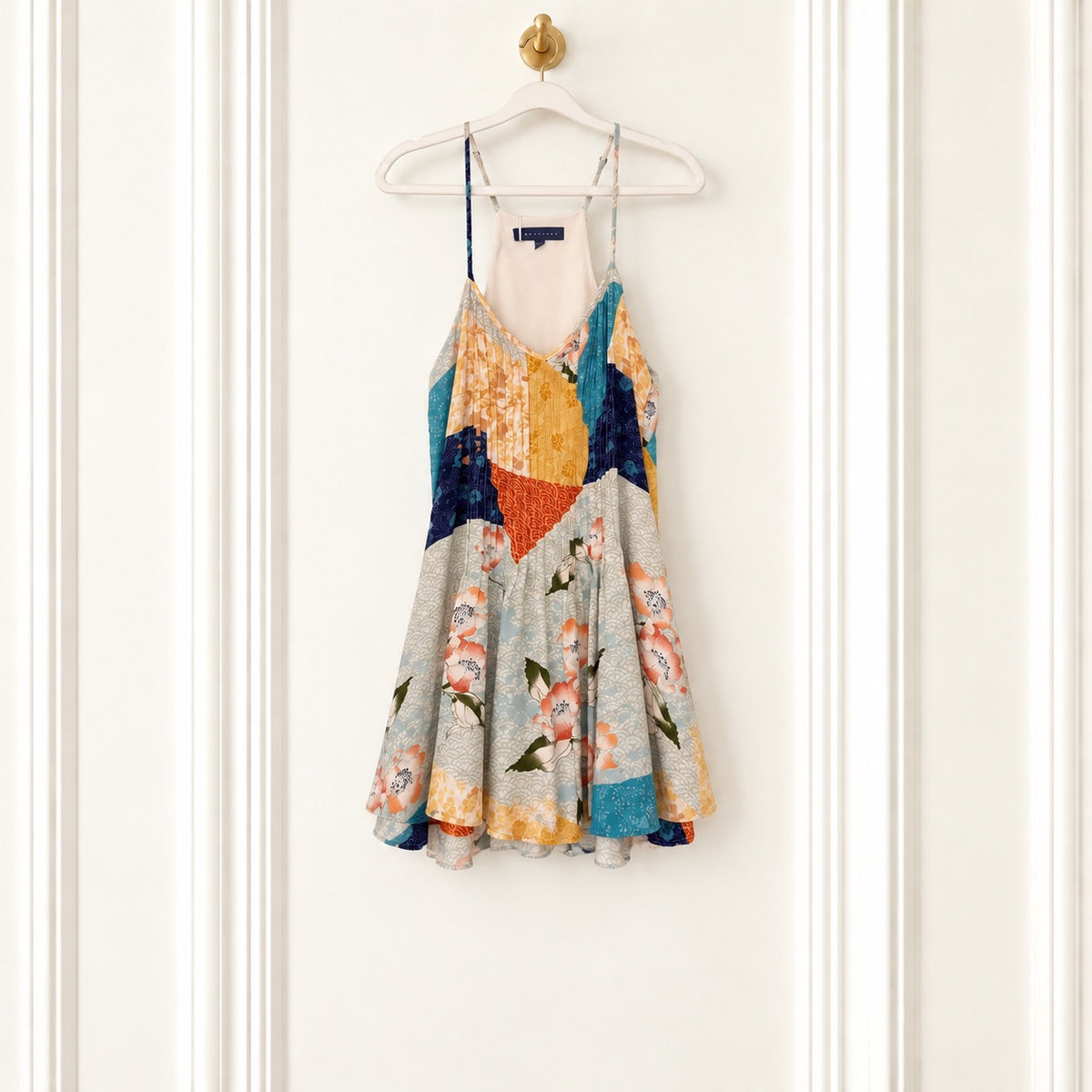 A sleeveless spaghetti-strap mini dress with a colorful abstract patchwork print, hanging on a white wooden hanger against a white paneled door.