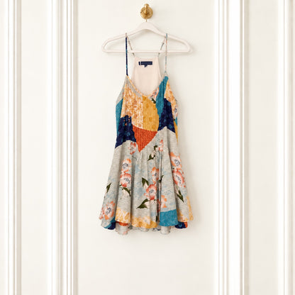 A sleeveless spaghetti-strap mini dress with a colorful abstract patchwork print, hanging on a white wooden hanger against a white paneled door.