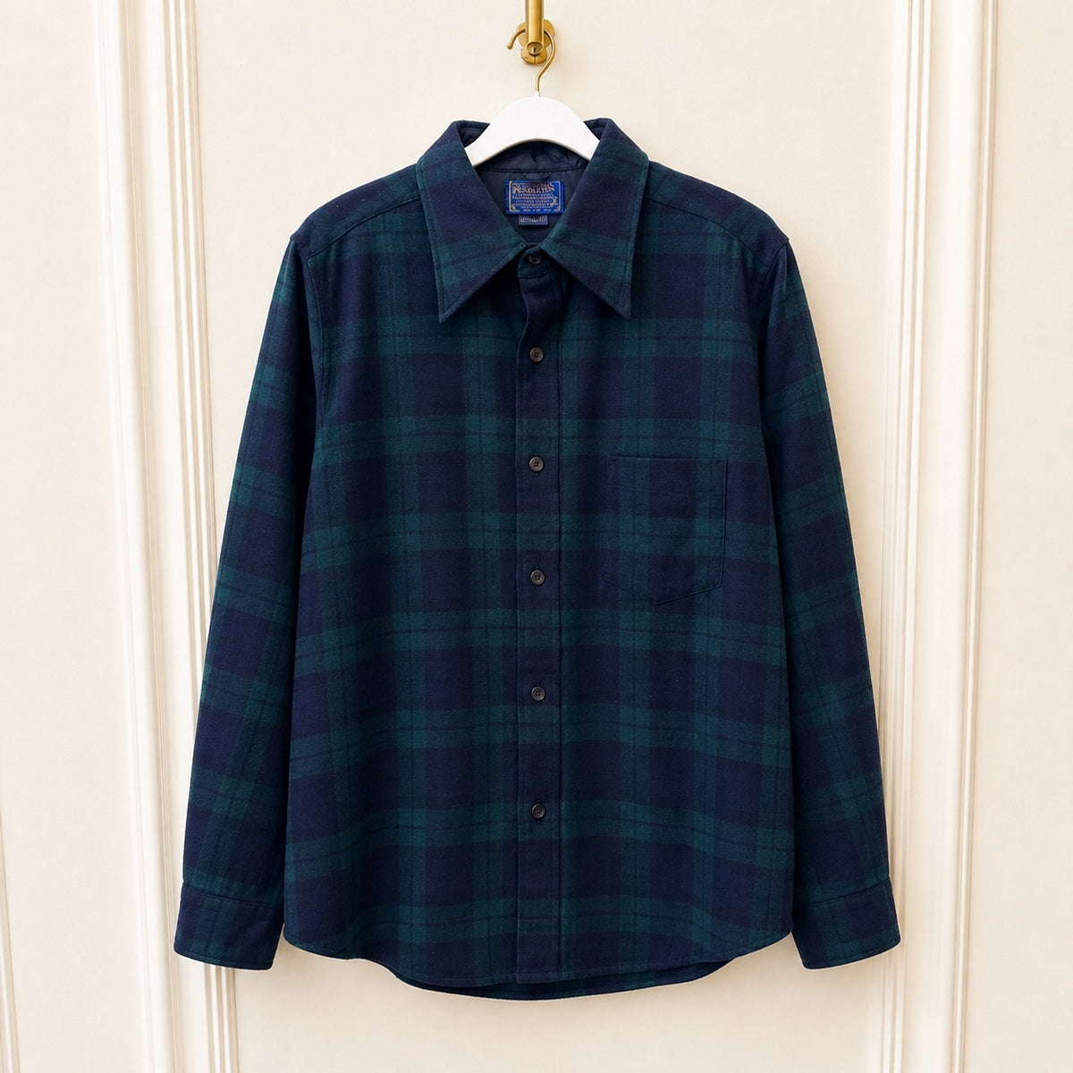 A long-sleeve button-up plaid shirt in dark navy and green, hanging on a white hanger against a cream paneled door.