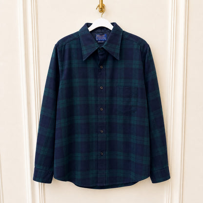 A long-sleeve button-up plaid shirt in dark navy and green, hanging on a white hanger against a cream paneled door.