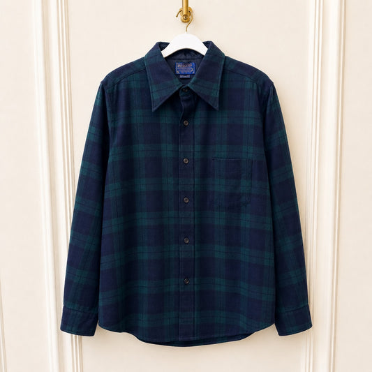 A long-sleeve button-up plaid shirt in dark navy and green, hanging on a white hanger against a cream paneled door.