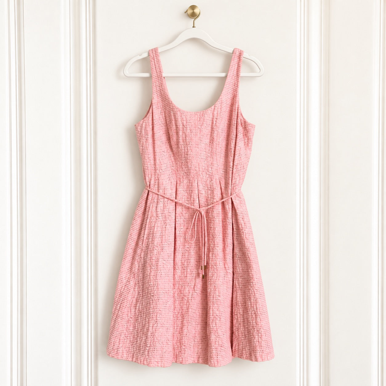 A pink textured sleeveless dress with a tie waist, hanging on a white wooden hanger against a white paneled door.