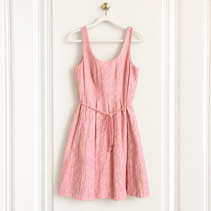A pink textured sleeveless dress with a tie waist, hanging on a white wooden hanger against a white paneled door.