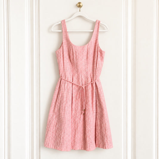 A pink textured sleeveless dress with a tie waist, hanging on a white wooden hanger against a white paneled door.