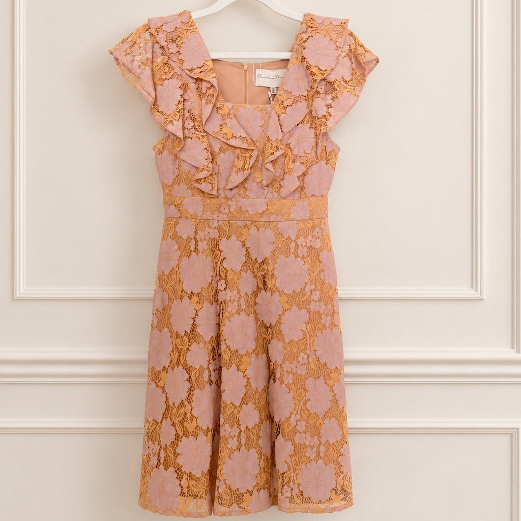 A sleeveless peach and pink floral lace dress with ruffled cap sleeves and a sweetheart neckline, hanging on a white hanger against a cream paneled wall.