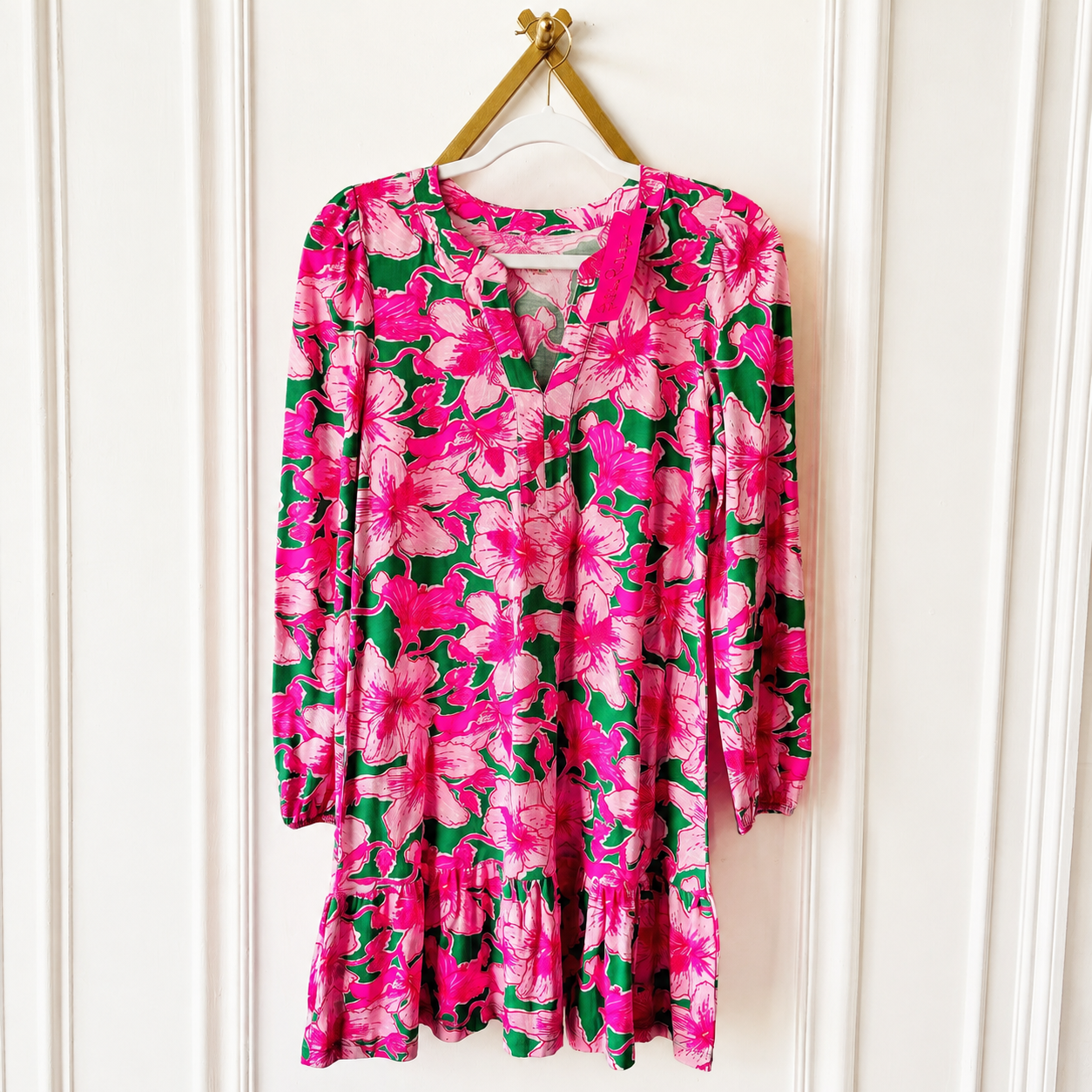 A long-sleeve floral dress with a vibrant pink and green pattern, hanging on a gold hanger against a white paneled door.