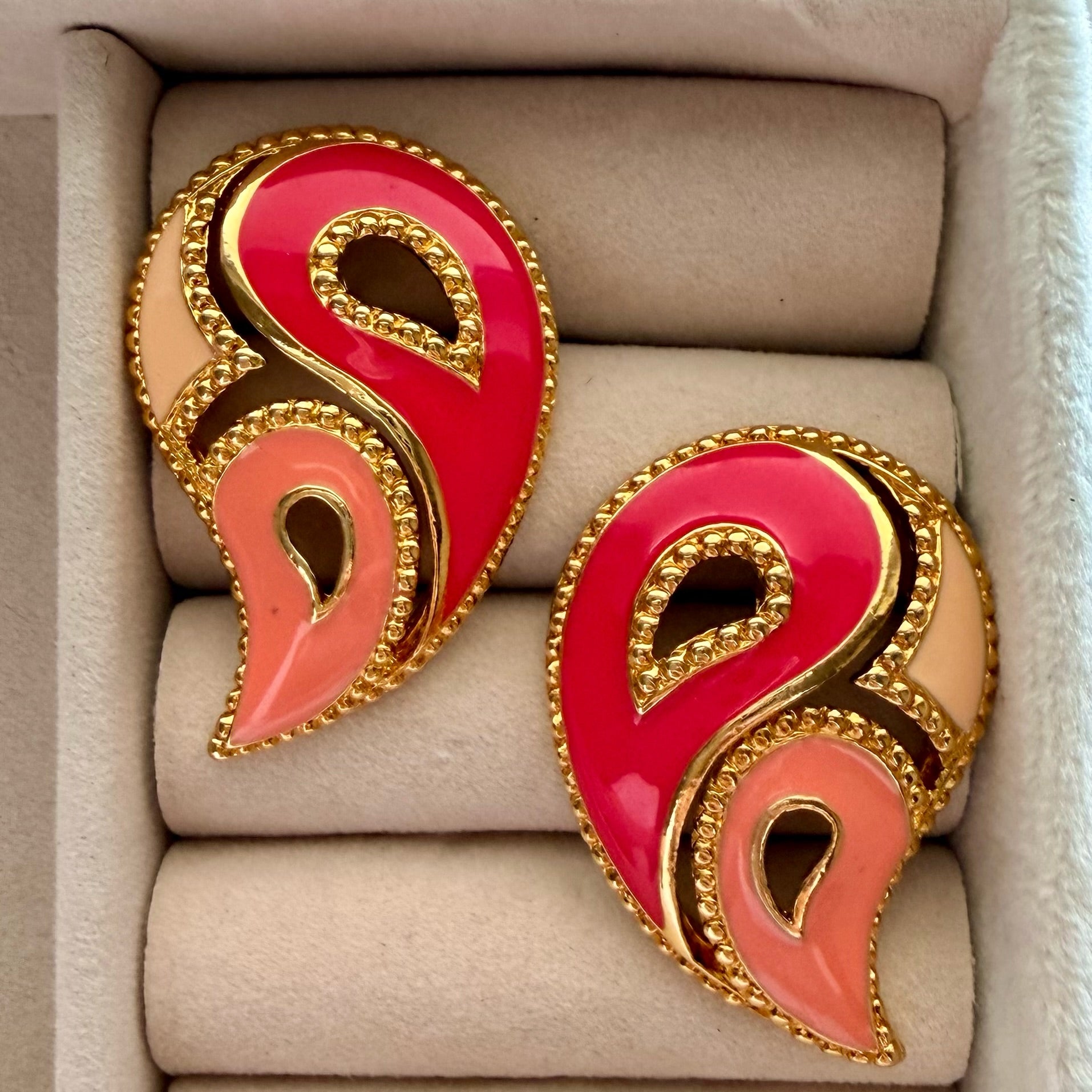 A pair of gold-tone paisley-shaped earrings with glossy pink and peach enamel, displayed in a beige jewelry box.
