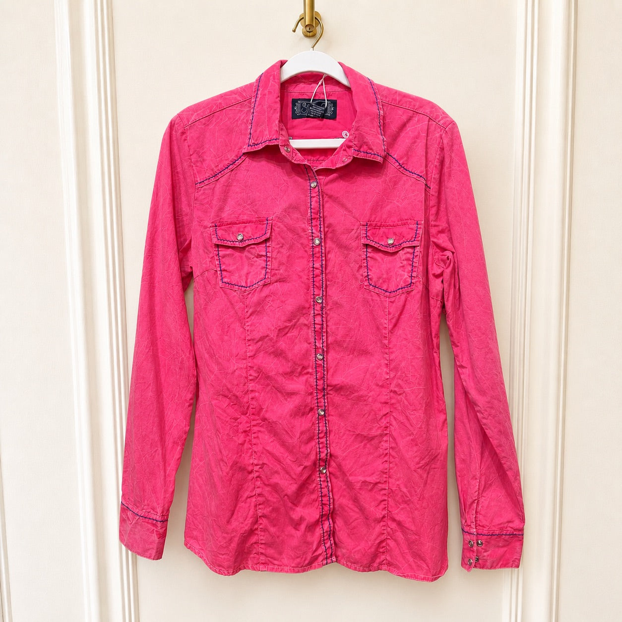 A bright pink western-style long-sleeve button-up shirt with contrast dark blue stitching, hanging on a white hanger against a cream-colored paneled door.