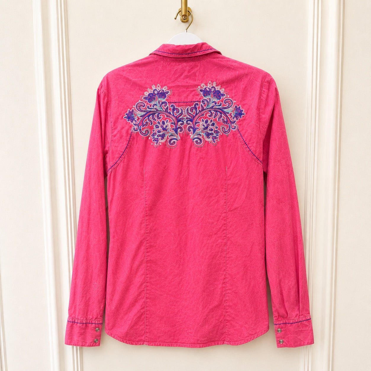 A bright pink long-sleeve button-up shirt hanging on a white hanger against a cream-colored paneled door. The back of the shirt features a prominent purple and blue floral embroidery across the upper back.