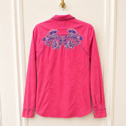 A bright pink long-sleeve button-up shirt hanging on a white hanger against a cream-colored paneled door. The back of the shirt features a prominent purple and blue floral embroidery across the upper back.