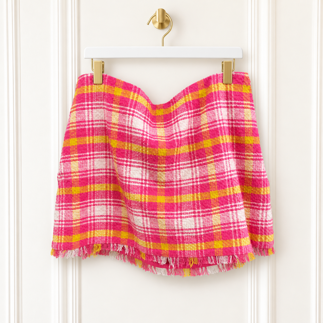 A pink and yellow plaid fringed mini skirt hanging from a white hanger with gold clips against a white paneled door.