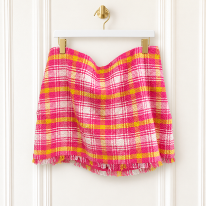 A pink and yellow plaid fringed mini skirt hanging from a white hanger with gold clips against a white paneled door.