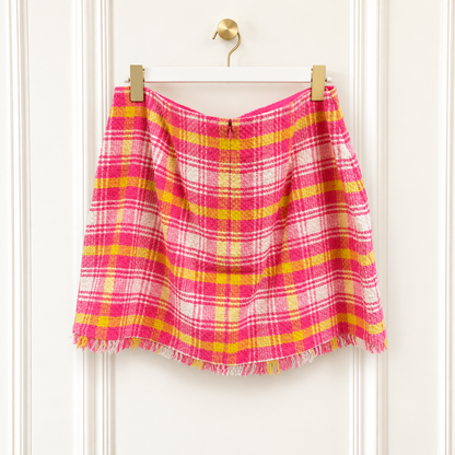 A pink and yellow plaid fringed skirt hanging from a white hanger with gold clips against a white paneled door.