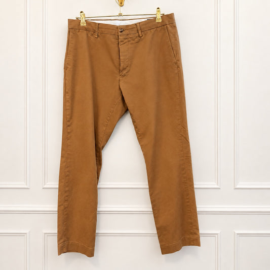 A pair of brown chino pants hanging from a gold hanger against a white paneled wall.