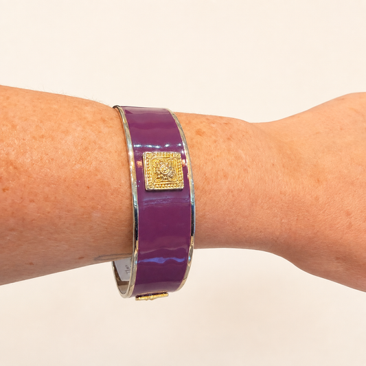 A close-up of a person's wrist wearing a glossy purple enamel cuff bracelet with a gold-toned square medallion clasp.