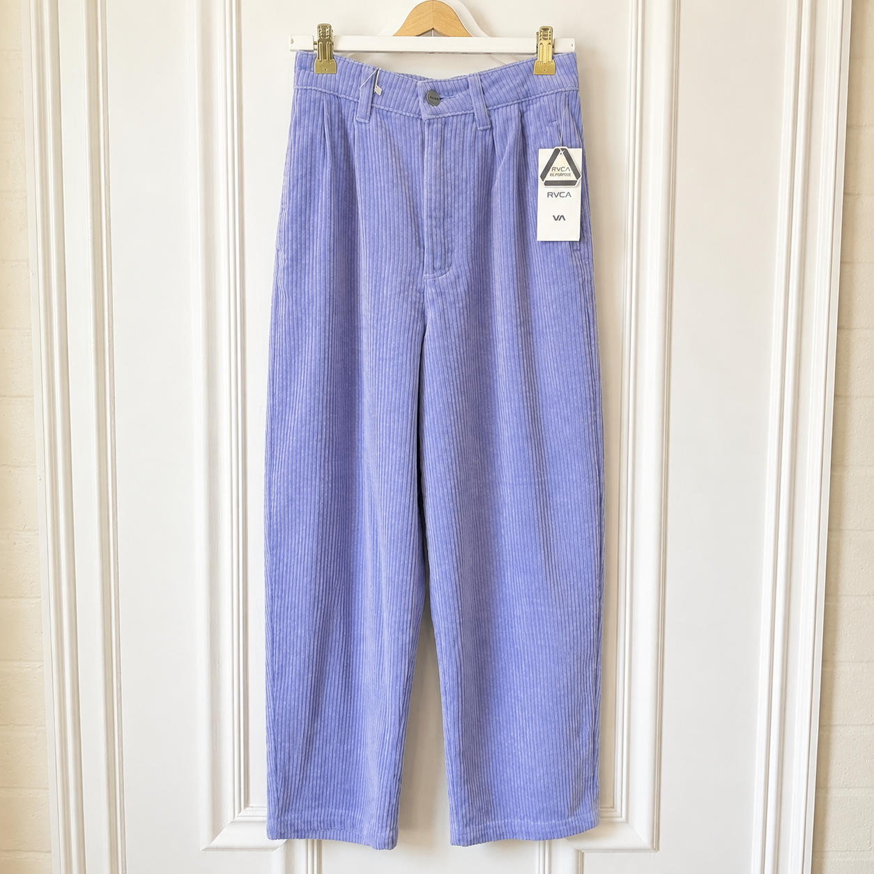 A pair of light purple corduroy cropped pants hanging on a wooden hanger against a white paneled door. A white paper tag is attached to the right pocket area.