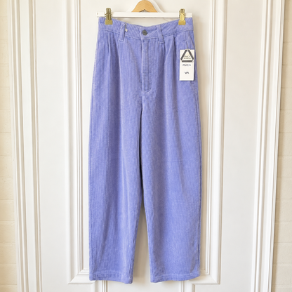 A pair of light purple corduroy cropped pants hanging on a wooden hanger against a white paneled door. A white paper tag is attached to the right pocket area.