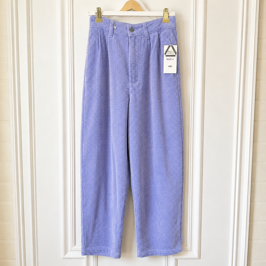 A pair of light purple corduroy cropped pants hanging on a wooden hanger against a white paneled door. A white paper tag is attached to the right pocket area.