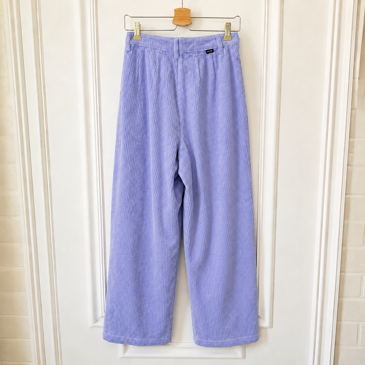 Light purple corduroy wide-leg cropped pants hanging on a wooden hanger against a white paneled door.
