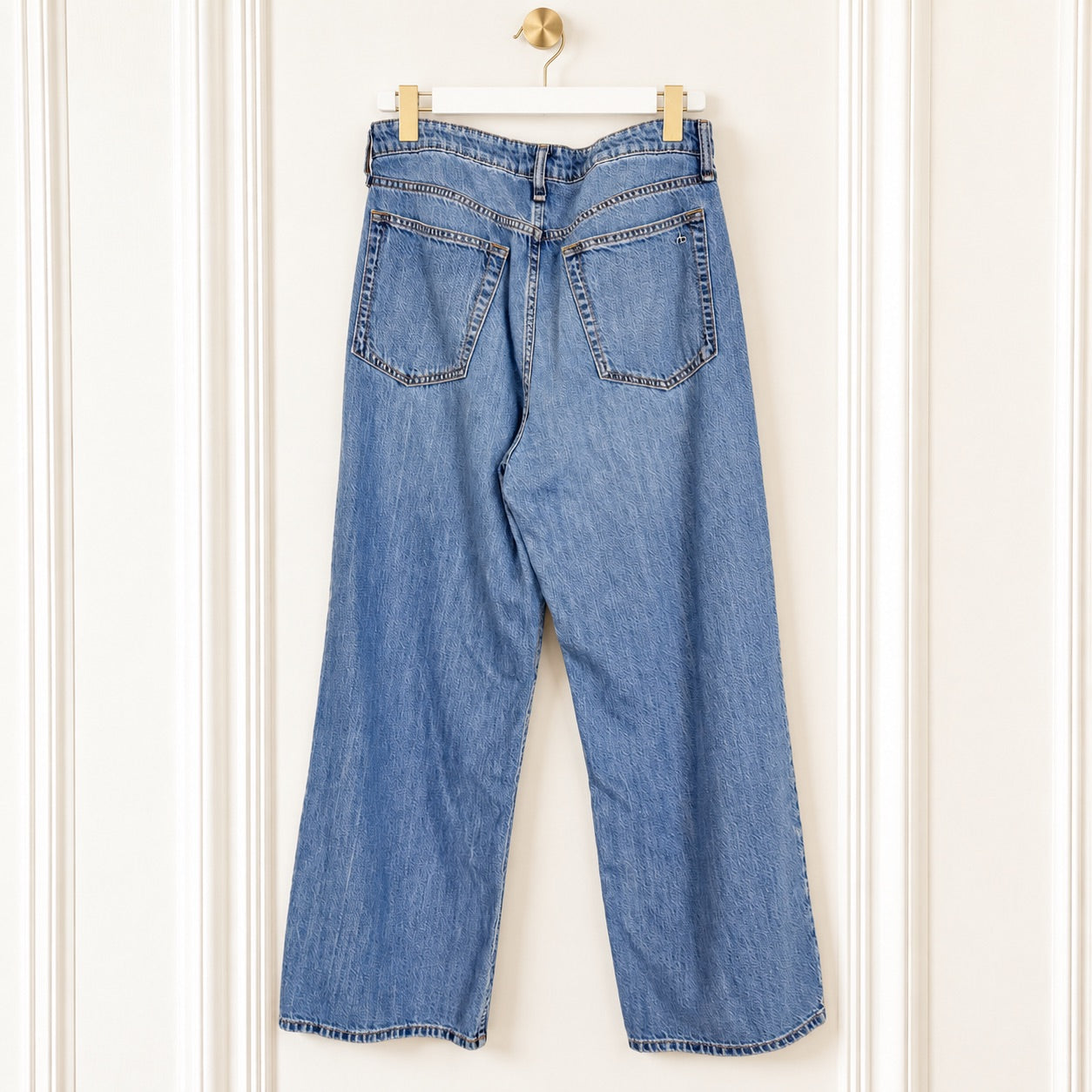 Light blue denim jeans shown from the back, hanging on a white hanger against a white paneled wall.