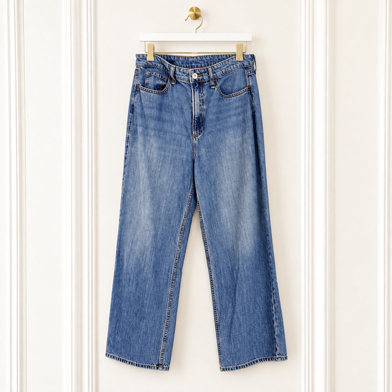 A pair of blue denim jeans with a straight-to-wide leg fit, hanging on a white wooden hanger against a white paneled wall.