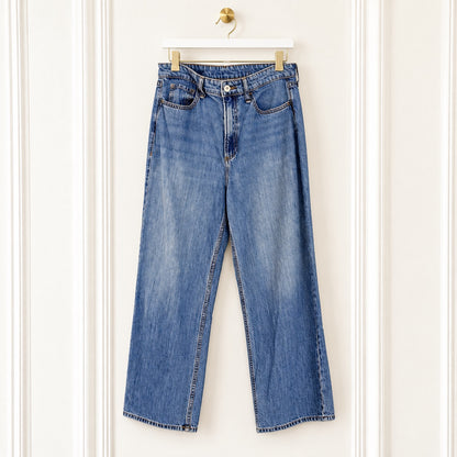 A pair of blue denim jeans with a straight-to-wide leg fit, hanging on a white wooden hanger against a white paneled wall.