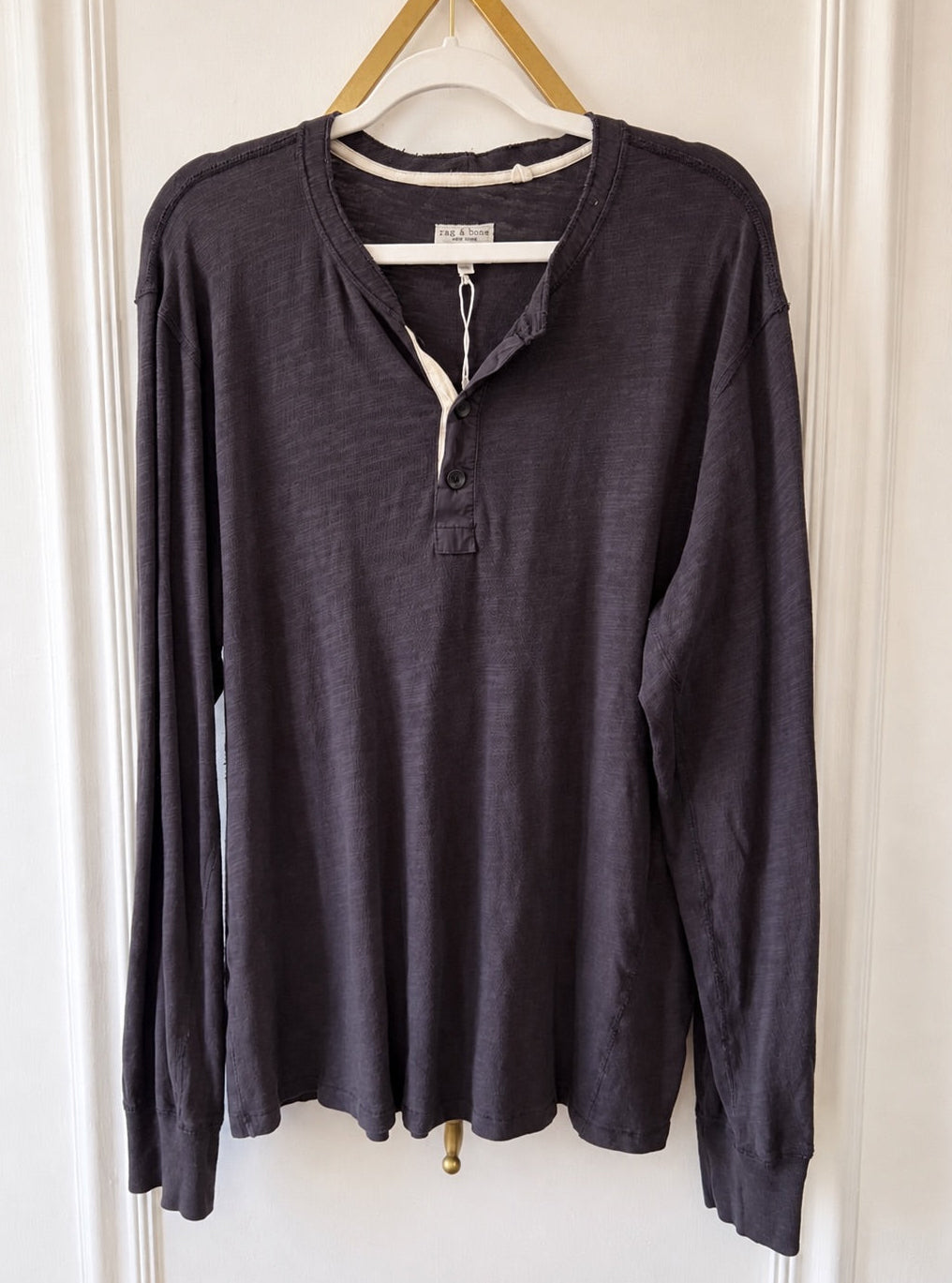 A dark charcoal gray long-sleeve henley shirt hanging on a wooden hanger against a white paneled door.