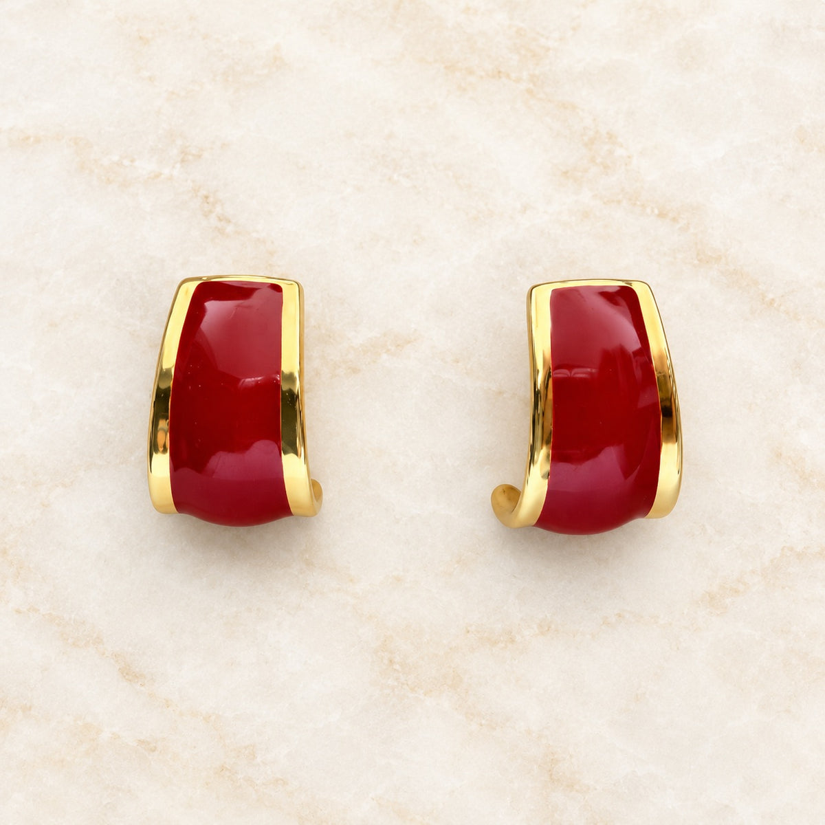 A pair of gold huggie hoop earrings with smooth, glossy deep red cabochon stones, displayed on a light beige marble surface.