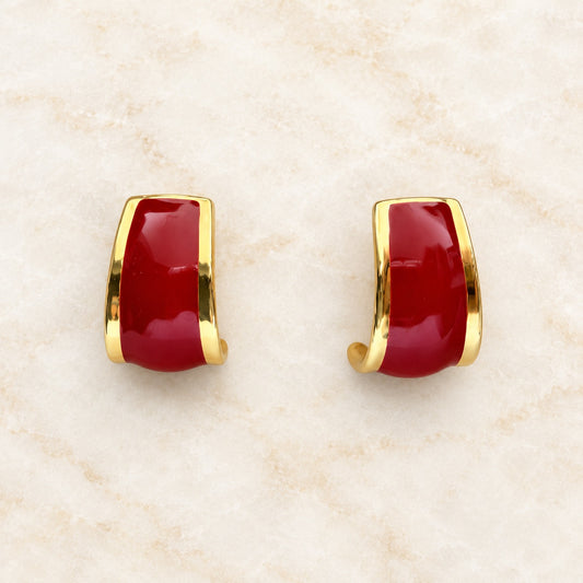 A pair of gold huggie hoop earrings with smooth, glossy deep red cabochon stones, displayed on a light beige marble surface.
