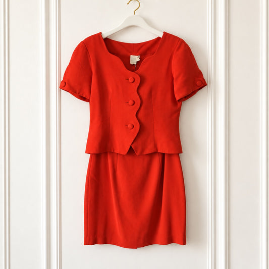 A bright red two-piece skirt suit set consisting of a short-sleeve peplum-style top with scalloped edges and button details, and a matching A-line skirt, hanging on a white hanger against a white paneled wall.