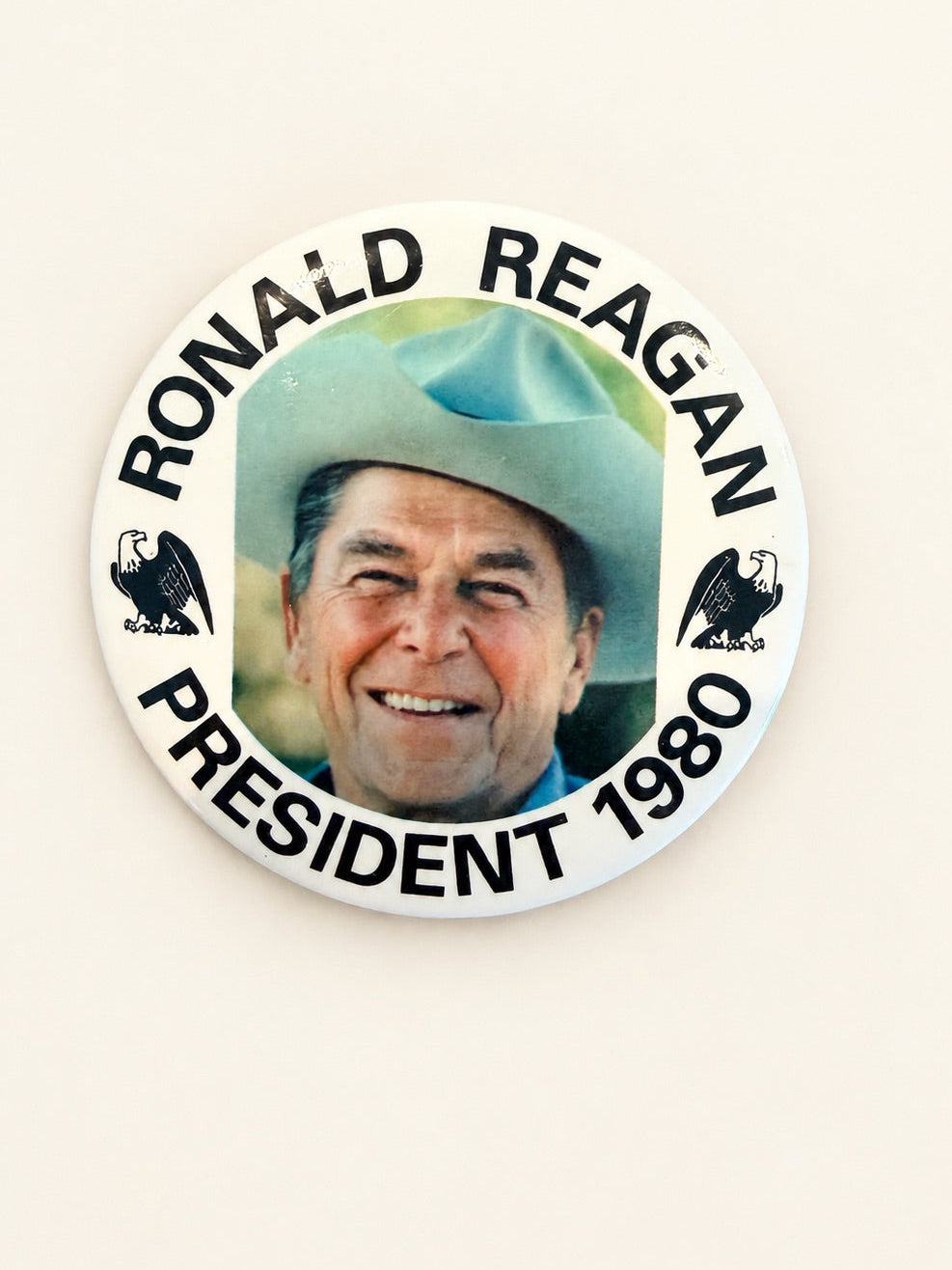 A round political button badge featuring a portrait of Ronald Reagan wearing a cowboy hat, with the text 'RONALD REAGAN PRESIDENT 1980' around the edge and eagle silhouettes on either side, shown against a plain off-white background.