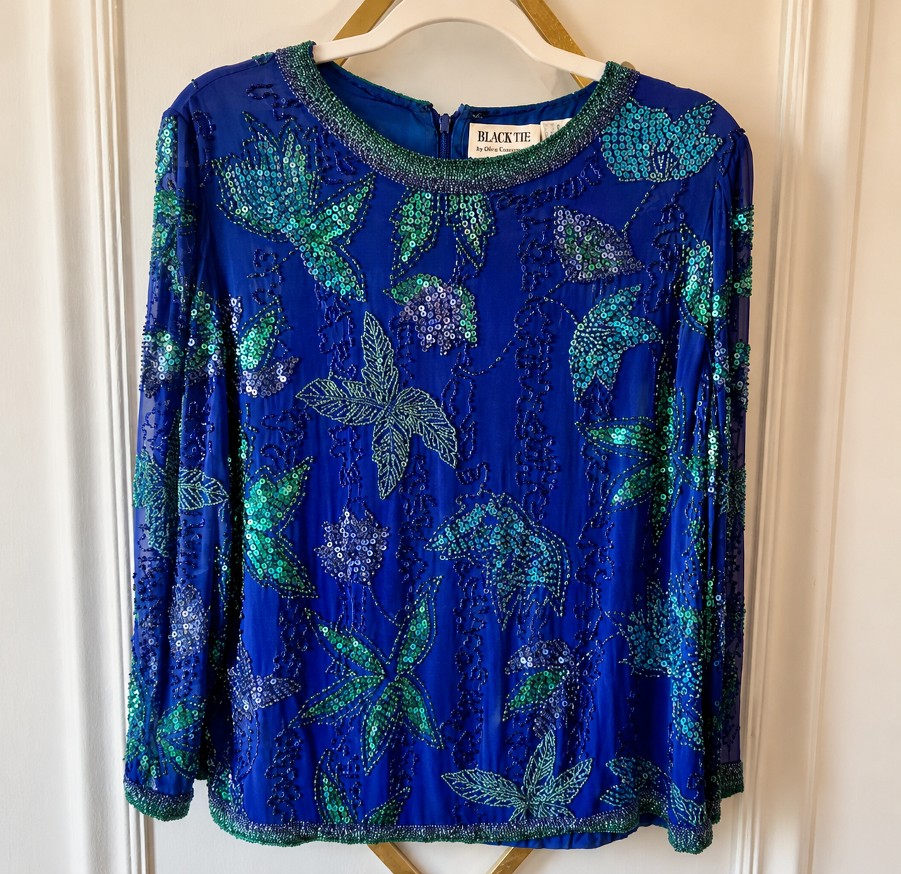 A royal blue long-sleeve blouse with green sequined floral and leaf patterns, hanging on a wooden hanger against a white paneled door.