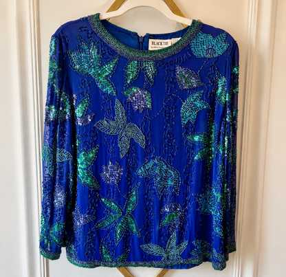 A royal blue long-sleeve blouse with green sequined floral and leaf patterns, hanging on a wooden hanger against a white paneled door.