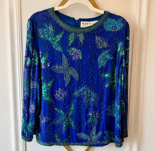 A royal blue long-sleeve blouse with green sequined floral and leaf patterns, hanging on a wooden hanger against a white paneled door.