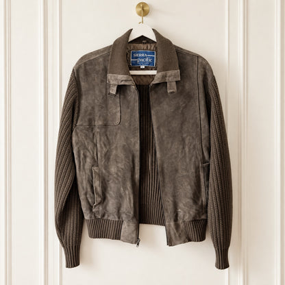 A dark brown suede jacket with ribbed knit sleeves and collar, hanging on a white hanger against a white paneled door. The brand label reads 'serpentine & Pacific'.