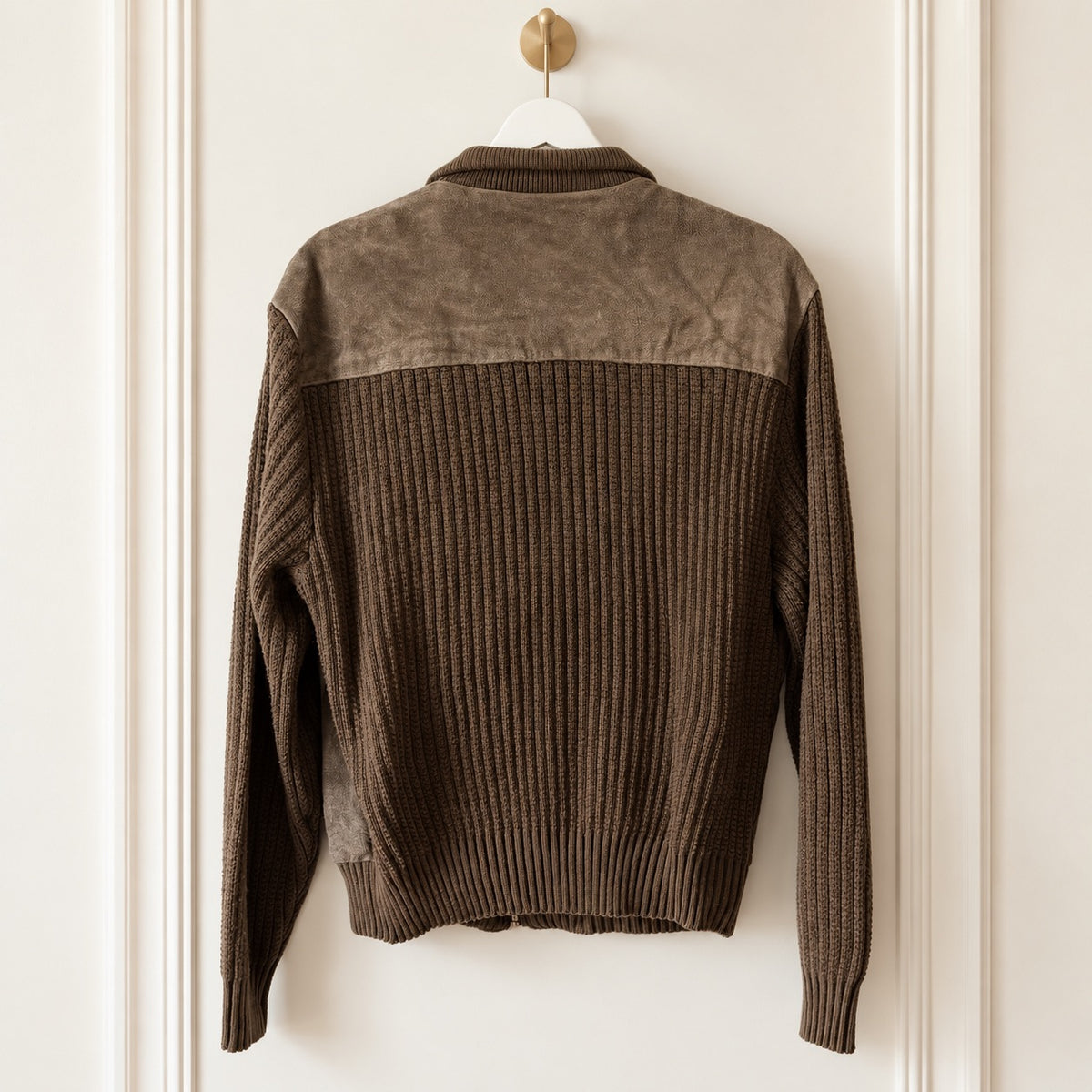 A brown color-block knit cardigan sweater with a suede-like shoulder yoke, hanging on a white hanger with a gold hook against a cream-colored paneled wall.