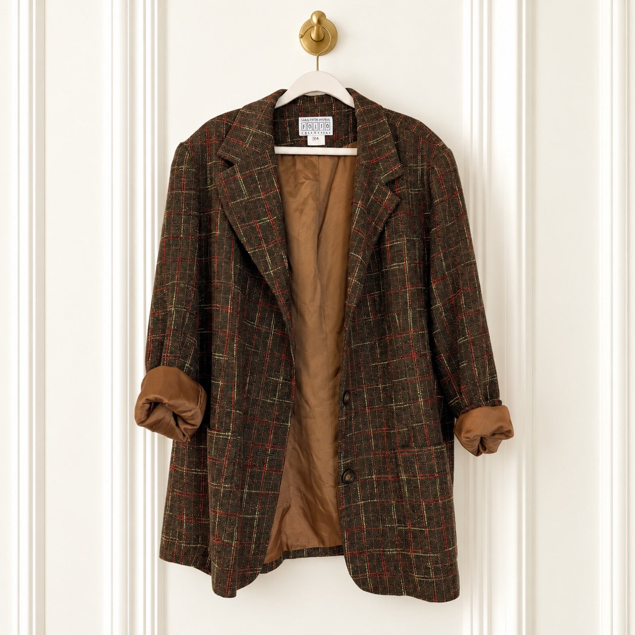 A brown plaid wool blazer with a tan lining, hanging on a white hanger with a gold ring pull against a white paneled door.