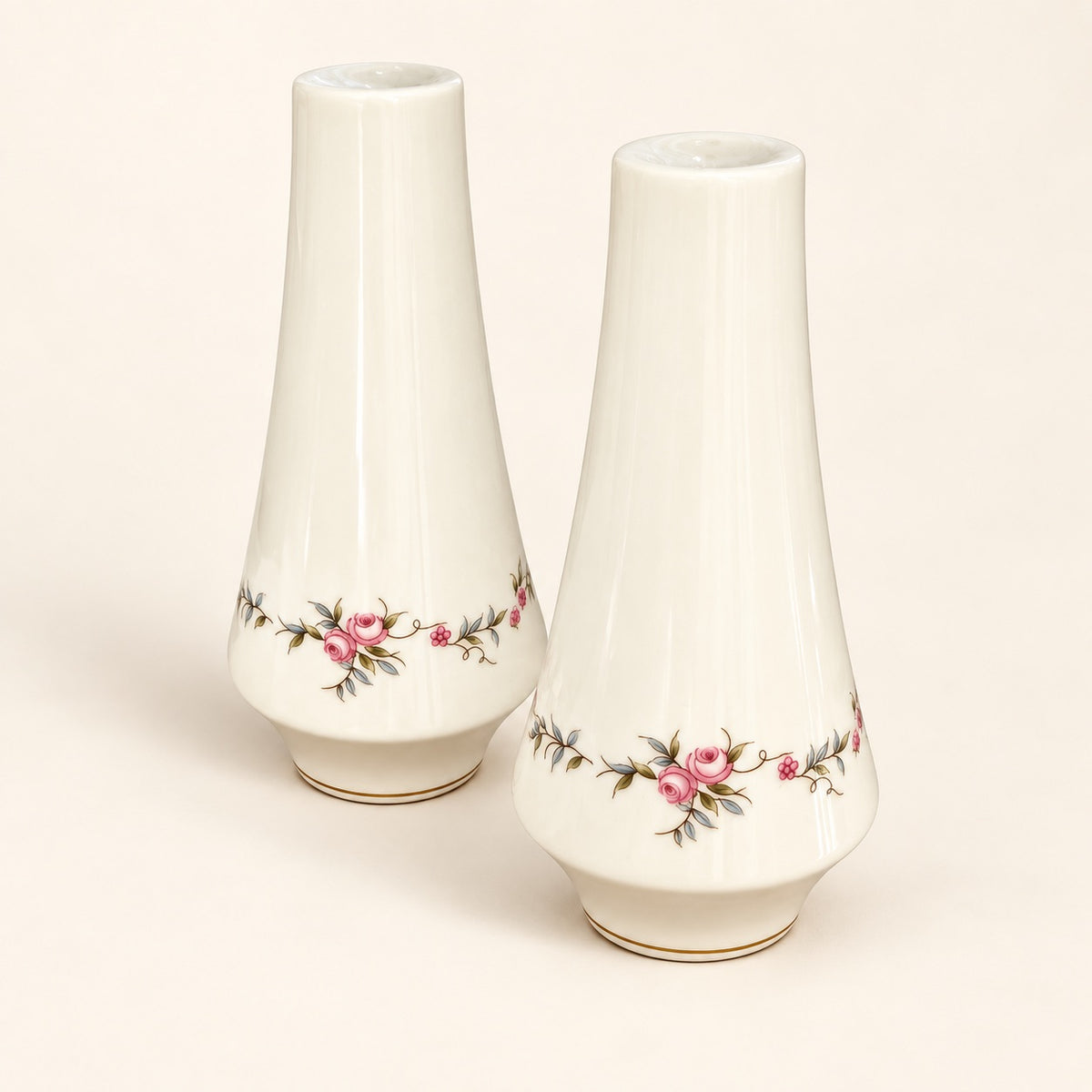 Two white ceramic vases with a delicate pink and green floral border design near the base, displayed on a warm cream background.