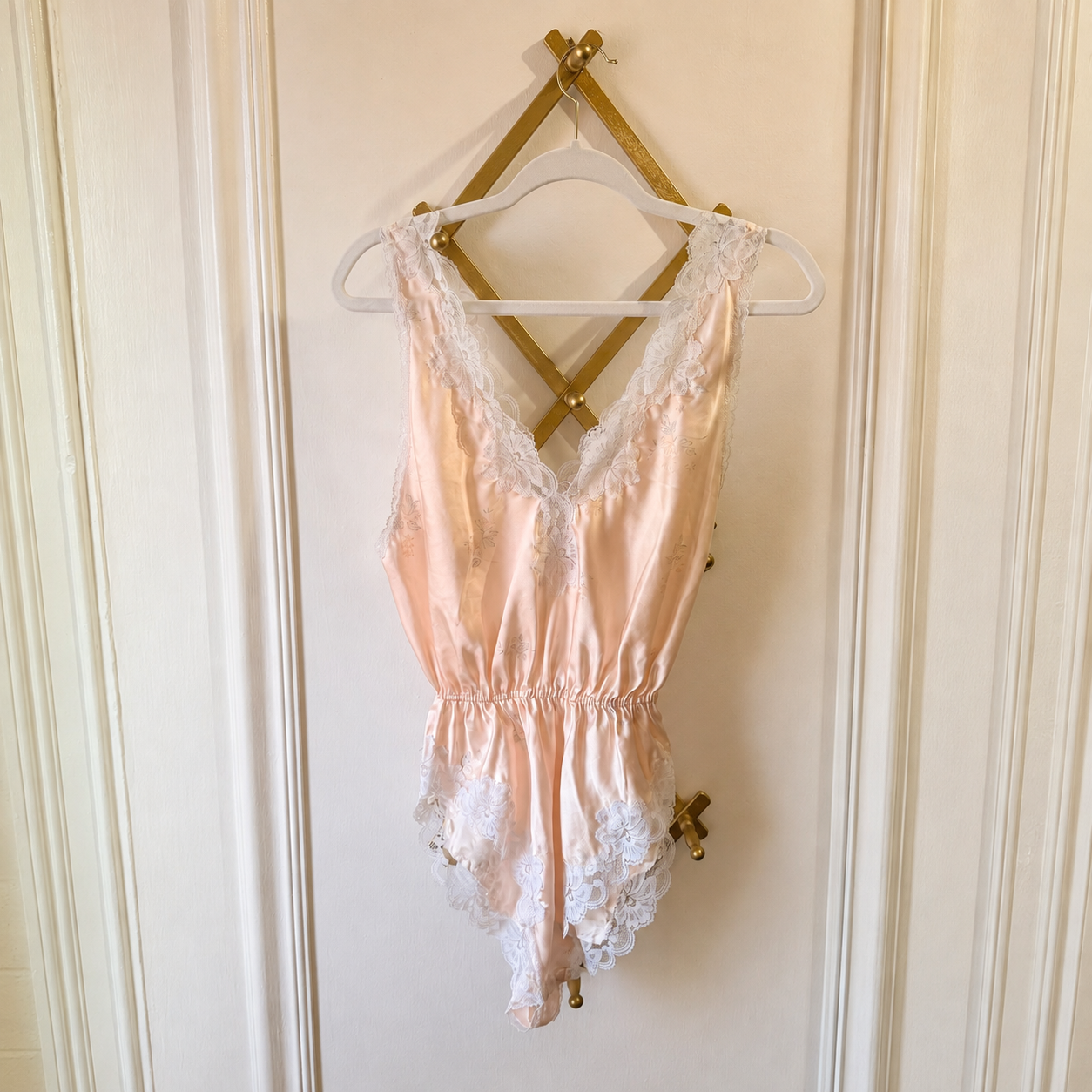 A pale peach satin babydoll lingerie piece with white lace trim, hanging on a white hanger from a gold wall hook against a cream paneled door.