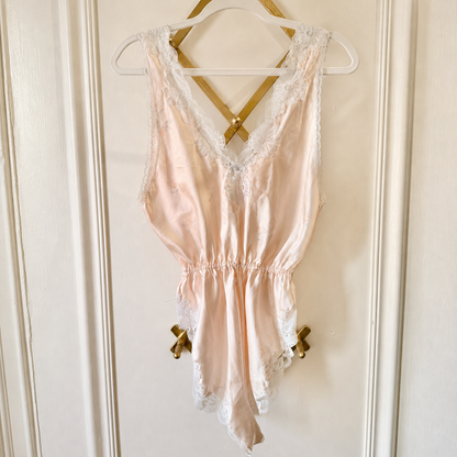 A pale blush pink satin babydoll lingerie chemise with white lace trim, hanging on a white hanger with gold ribbon bows against a cream-colored paneled door.