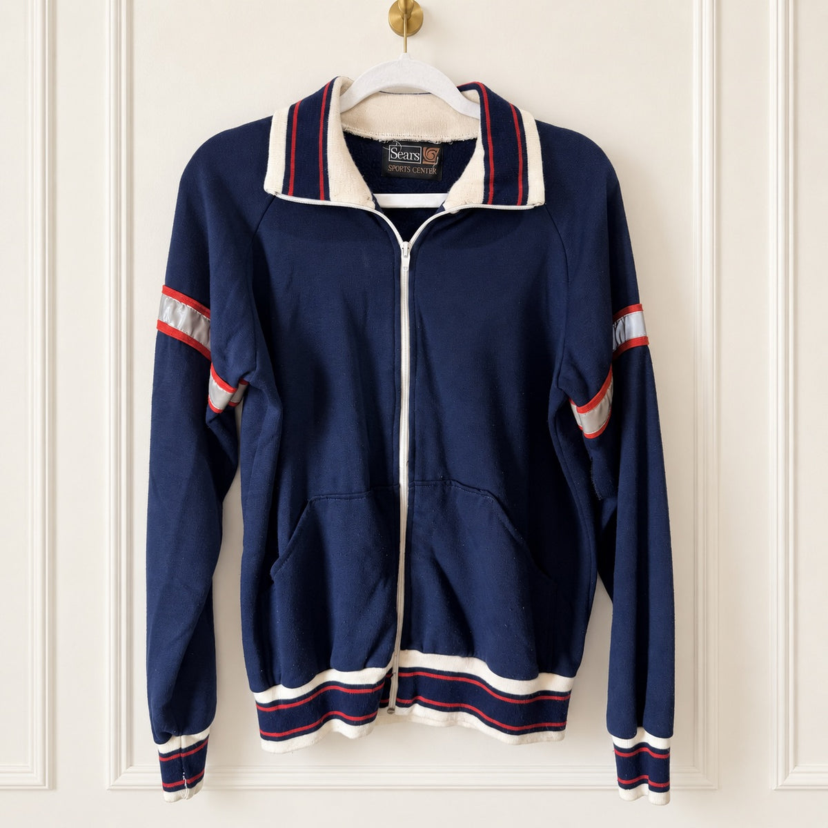 A vintage navy blue zip-up track jacket with white and red striped ribbed trim, hanging on a white hanger against a white paneled door.
