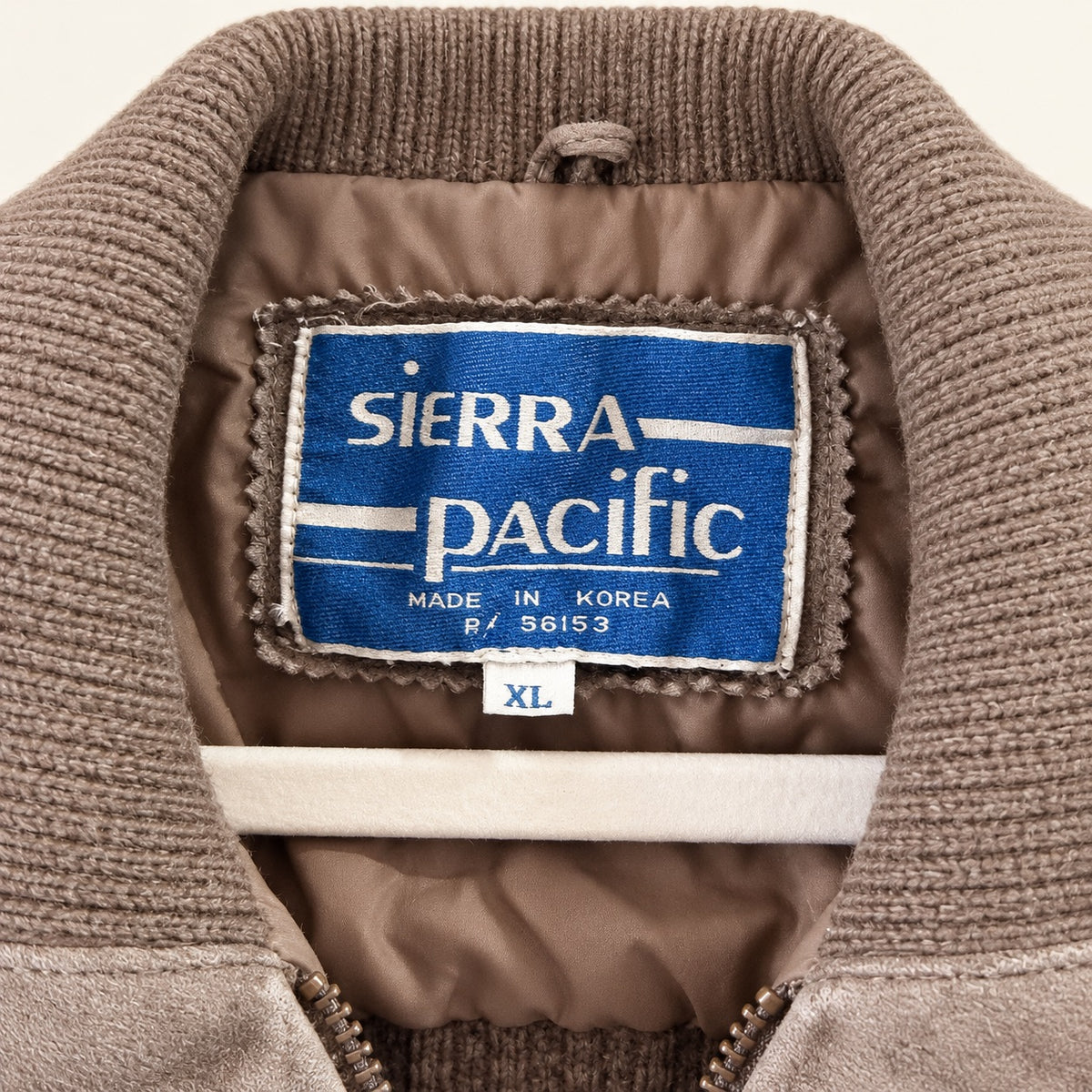 Close-up of the interior collar and neck label of a brown ribbed knit jacket. The blue and white woven label reads 'SIERRA PACIFIC' with 'MADE IN KOREA' and a small size tag reading 'XL'.