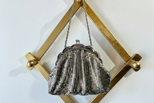 A vintage-style silver mesh clutch purse with a scalloped metal frame and chain strap, hanging from a gold-colored geometric wall rack against a white wall.