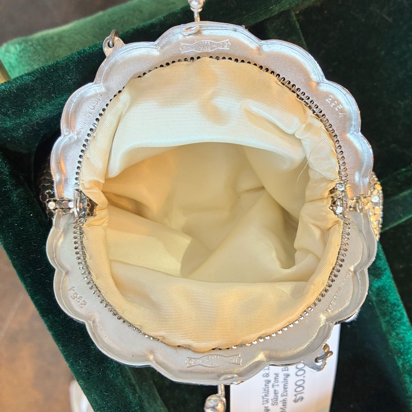 A vintage silver-toned purse with a scalloped edge and cream satin lining, displayed inside a green velvet box with a price tag hanging from it.