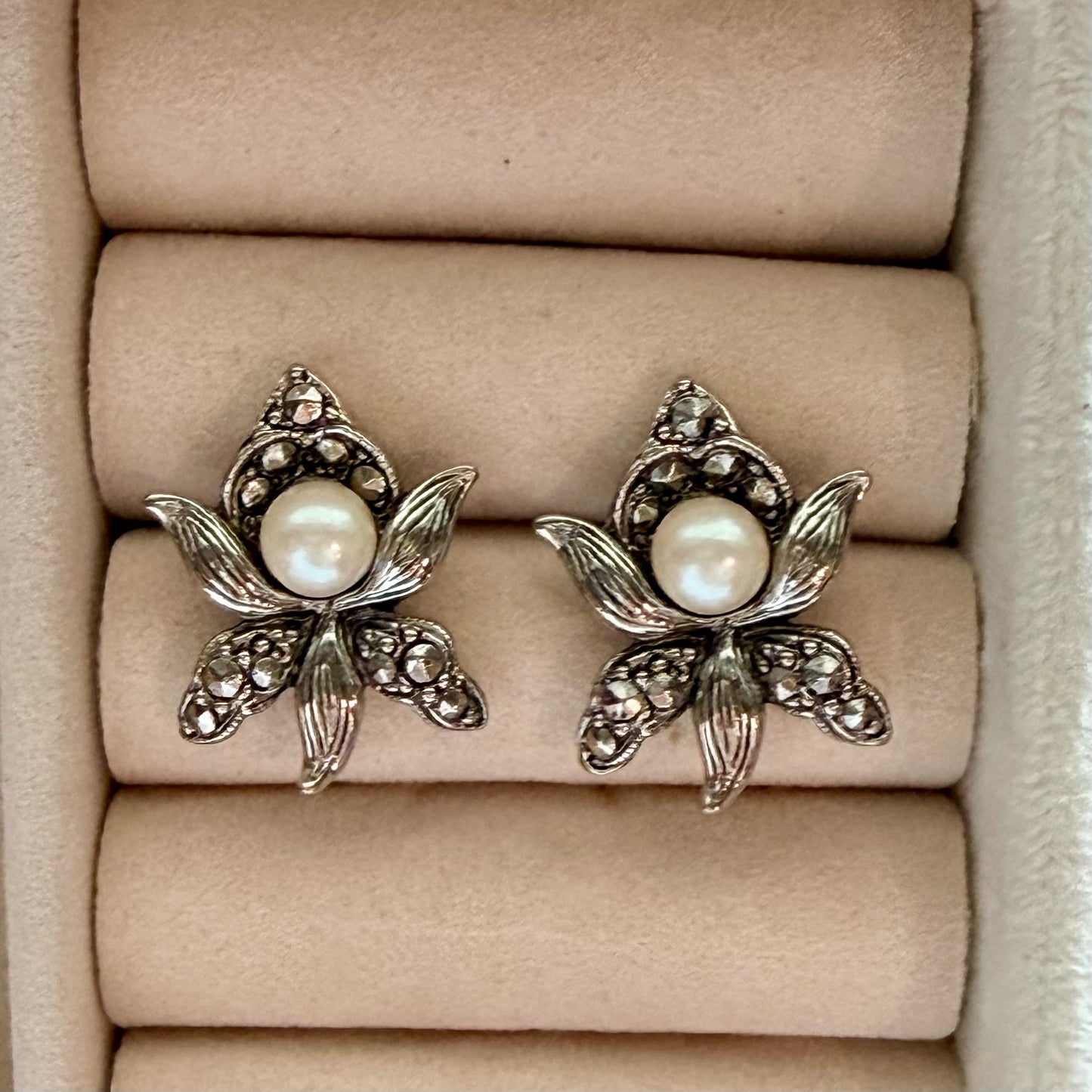 A pair of silver-toned flower-shaped earrings with white pearl centers and small clear stones, displayed on a beige jewelry box cushion.