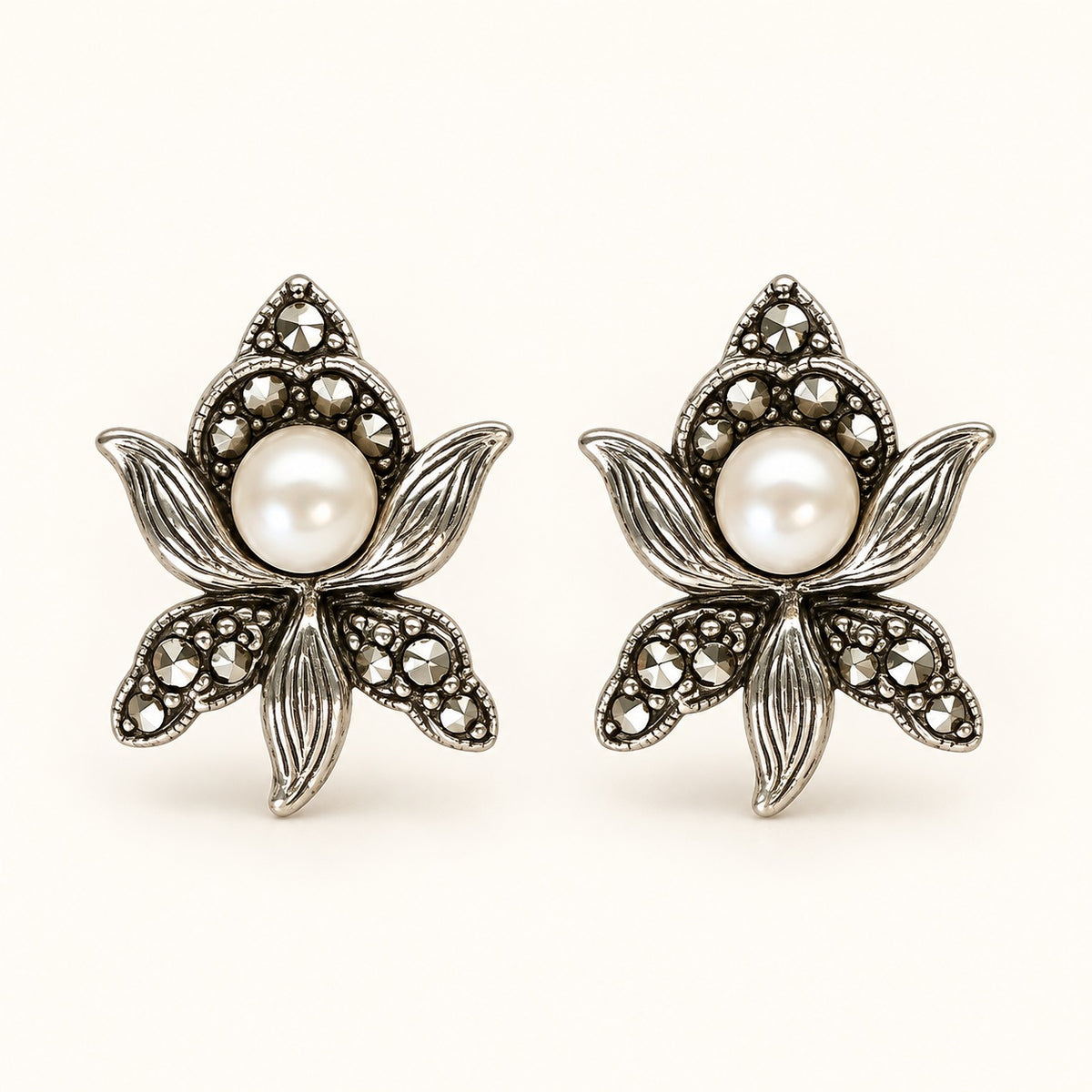 A pair of silver-toned floral stud earrings featuring a central white pearl surrounded by small clear stones, shown on a plain light beige background.
