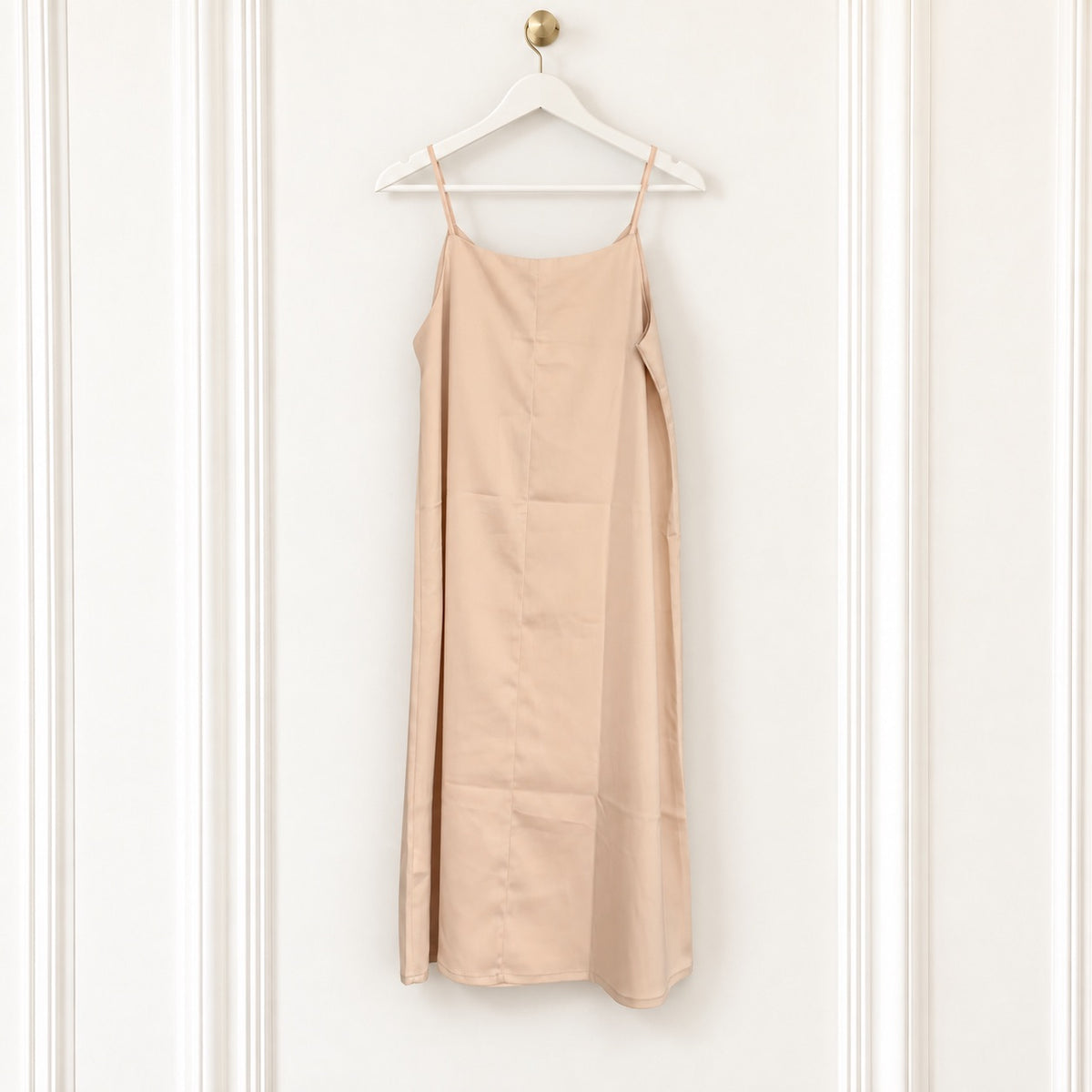 A light beige slip dress with thin spaghetti straps hanging from a white wooden hanger on a white paneled wall.