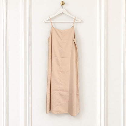 A light beige slip dress with thin spaghetti straps hanging from a white wooden hanger on a white paneled wall.