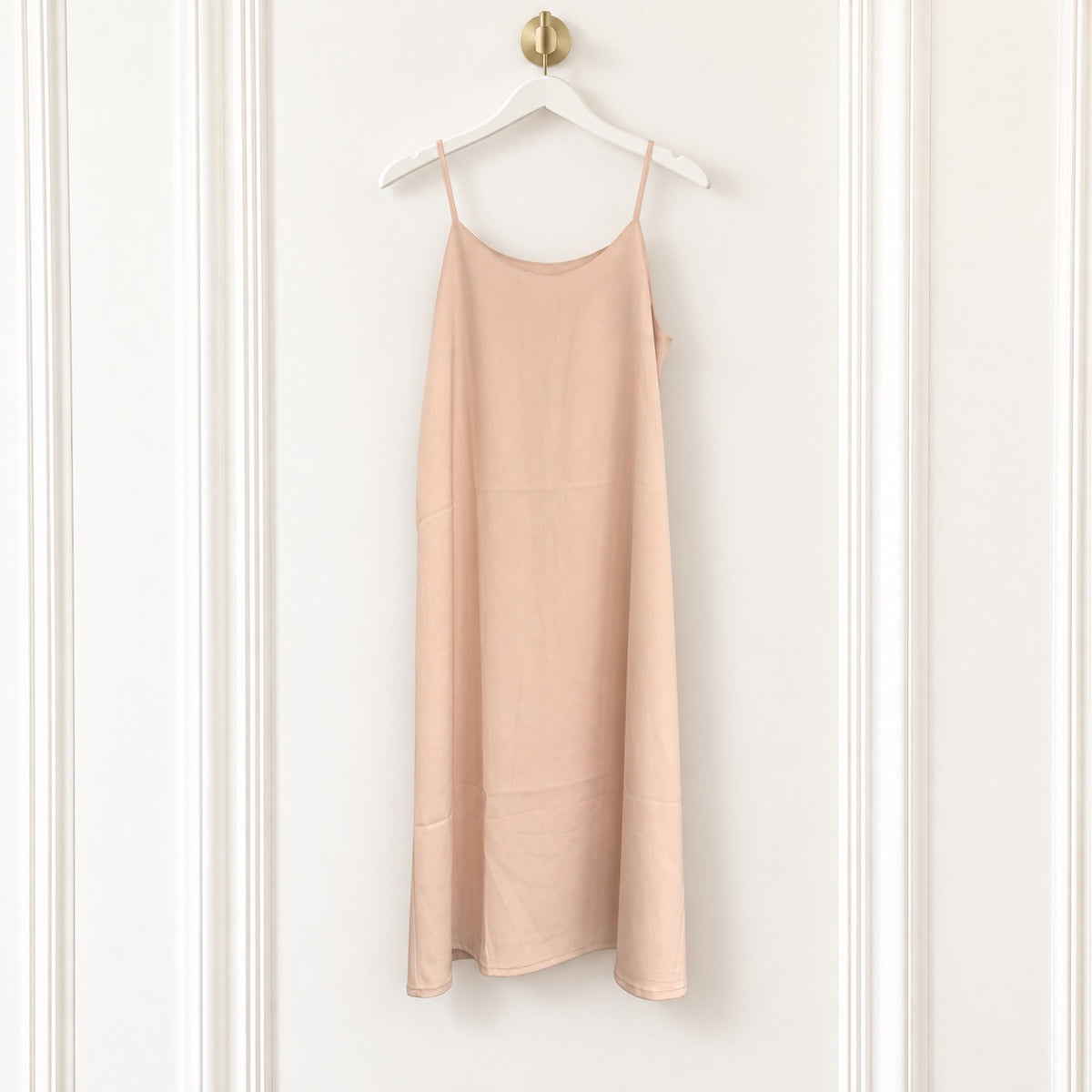 A light peach-pink linen slip dress with thin spaghetti straps, hanging on a white wooden hanger with a gold knob against a white paneled wall.