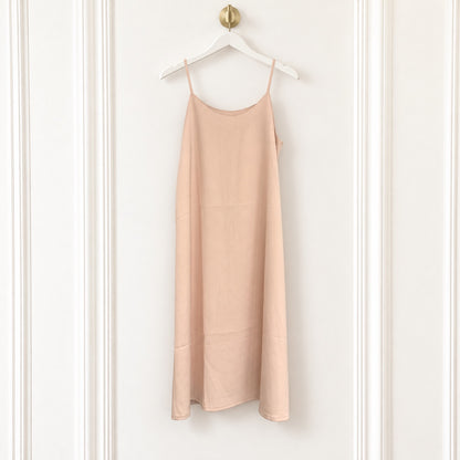 A light peach-pink linen slip dress with thin spaghetti straps, hanging on a white wooden hanger with a gold knob against a white paneled wall.